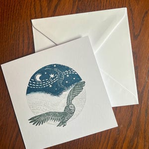 May include: A white greeting card with a teal and gray owl illustration, a crescent moon, and stars. The card is accompanied by a white envelope, set against a dark wood surface. The owl is depicted in flight.