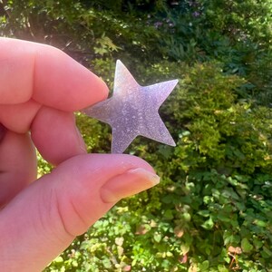 May include: A small, iridescent, five-pointed star made of glass or plastic. The star is held in a hand against a blurred background of green foliage.