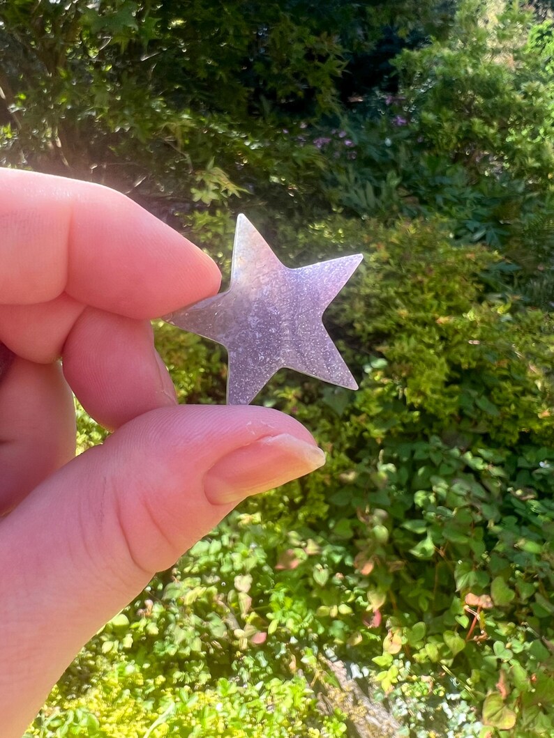 May include: A small, iridescent, five-pointed star made of glass or plastic. The star is held in a hand against a blurred background of green foliage.