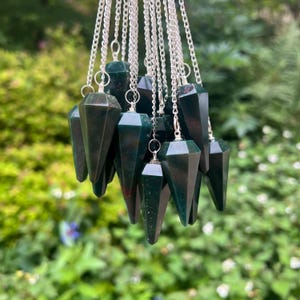 May include: A group of dark green gemstone pendulums suspended from silver chains. Each pendulum is faceted and pointed. The gemstones display a mottled pattern with varying shades of green. The background is a blurred view of green foliage.