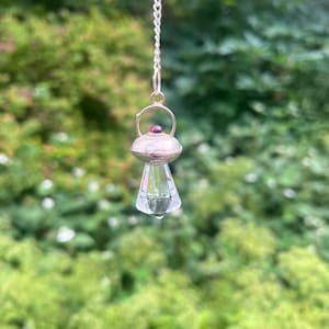 May include: A silver-toned pendant hanging from a chain, featuring a metallic UFO-shaped cap with a small embedded magenta gemstone. Below the cap, a clear, faceted, conical crystal point extends downwards, catching the light.