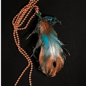 Puede incluir: Un collar de cadena de cobre con un colgante de pluma de color turquesa y cobre. La pluma tiene un agujero en el centro y est&aacute; unida a la cadena con un lazo de cobre.