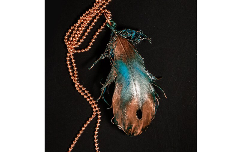 Puede incluir: Un collar de cadena de cobre con un colgante de pluma de color turquesa y cobre. La pluma tiene un agujero en el centro y est&aacute; unida a la cadena con un lazo de cobre.