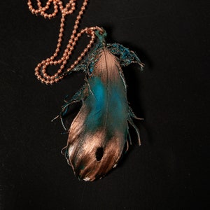 Puede incluir: Un collar de cadena de cobre con un colgante de pluma de color turquesa y cobre. La pluma est&aacute; detallada con una superficie texturizada y tiene un agujero en el centro.