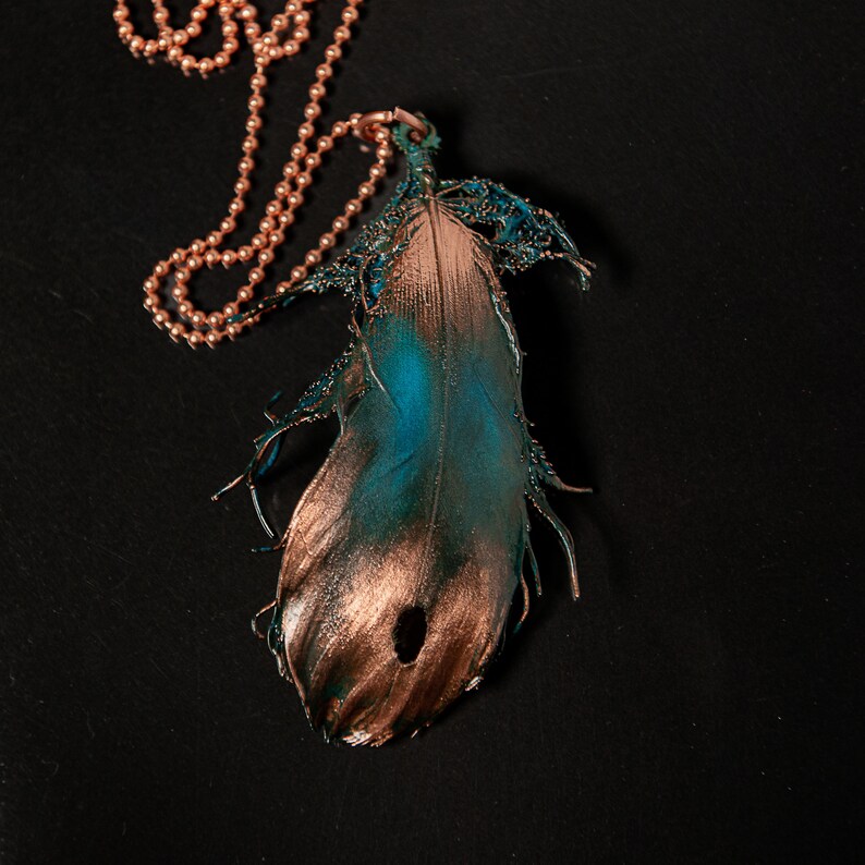 Puede incluir: Un collar de cadena de cobre con un colgante de pluma de color turquesa y cobre. La pluma est&aacute; detallada con una superficie texturizada y tiene un agujero en el centro.