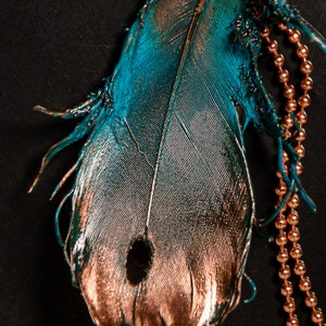 Puede incluir: Una sola pluma de color turquesa y cobre con un agujero en el centro. La pluma est&aacute; unida a una cadena de cobre.