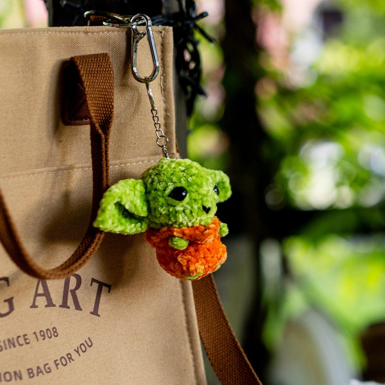 Puede incluir: Un bolso de tela beige con correas marrones y el texto "G ART SINCE 1908 ON BAG FOR YOU" impreso en el frente. Unido al bolso hay un peque&ntilde;o llavero de peluche verde tejido a ganchillo que representa una criatura con una prenda naranja.