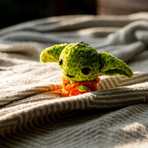 Puede incluir: Un peque&ntilde;o juguete de peluche tejido a mano con una cabeza de color verde brillante, orejas grandes y puntiagudas y ojos oscuros y redondos. La figura lleva una prenda texturizada de color naranja alrededor de la cintura y tiene una anilla met&aacute;lica.