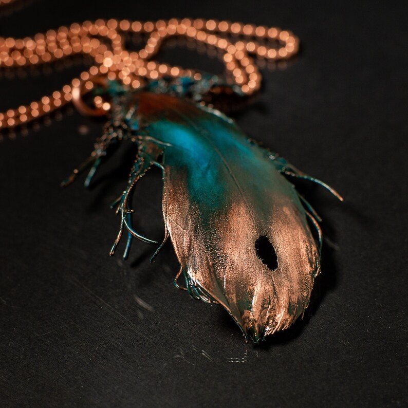 Puede incluir: Un collar de cobre con un colgante de pluma. La pluma est&aacute; pintada con una p&aacute;tina turquesa y cobre.