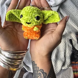 Puede incluir: Un peque&ntilde;o llavero de ganchillo hecho a mano que representa un personaje con cabeza de color verde lima brillante, grandes ojos de pl&aacute;stico negro y orejas anchas. El cuerpo tiene una textura de hilo naranja vibrante y un anillo de metal plateado.