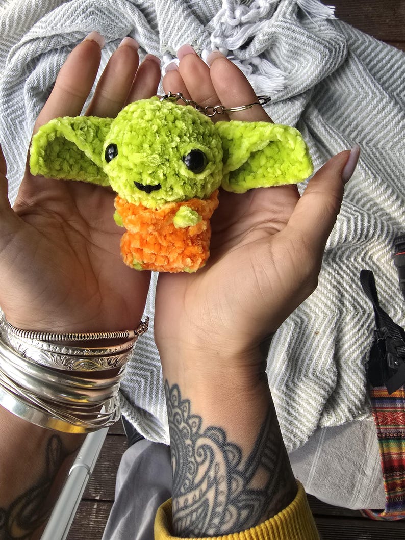 Puede incluir: Un peque&ntilde;o llavero de ganchillo hecho a mano que representa un personaje con cabeza de color verde lima brillante, grandes ojos de pl&aacute;stico negro y orejas anchas. El cuerpo tiene una textura de hilo naranja vibrante y un anillo de metal plateado.