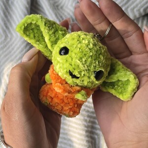 Puede incluir: Un peque&ntilde;o llavero de ganchillo hecho a mano que representa una criatura verde con grandes orejas puntiagudas y ojos de pl&aacute;stico negro. La figura lleva una prenda texturizada de color naranja y est&aacute; unida a una anilla met&aacute;lica plateada.