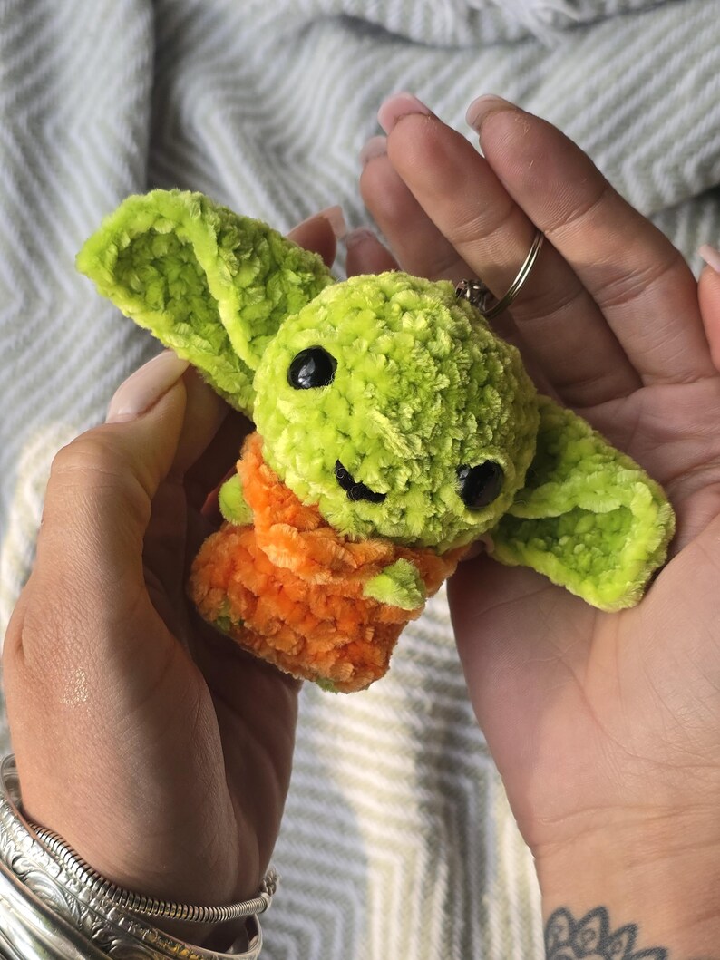 Puede incluir: Un peque&ntilde;o llavero de ganchillo hecho a mano que representa una criatura verde con grandes orejas puntiagudas y ojos de pl&aacute;stico negro. La figura lleva una prenda texturizada de color naranja y est&aacute; unida a una anilla met&aacute;lica plateada.