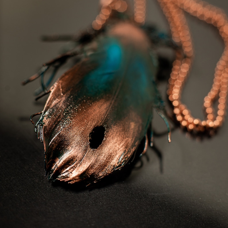 Puede incluir: Un colgante de pluma de cobre y turquesa en una cadena de cobre. La pluma tiene un agujero en el centro y est&aacute; detallada con un acabado texturizado.