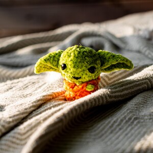 Puede incluir: Un peque&ntilde;o peluche de ganchillo hecho a mano con una cabeza redonda verde, ojos grandes, oscuros y brillantes, y orejas anchas y puntiagudas. El cuerpo est&aacute; texturizado con hilo naranja brillante, dando un aspecto suave y esponjoso a la figura.