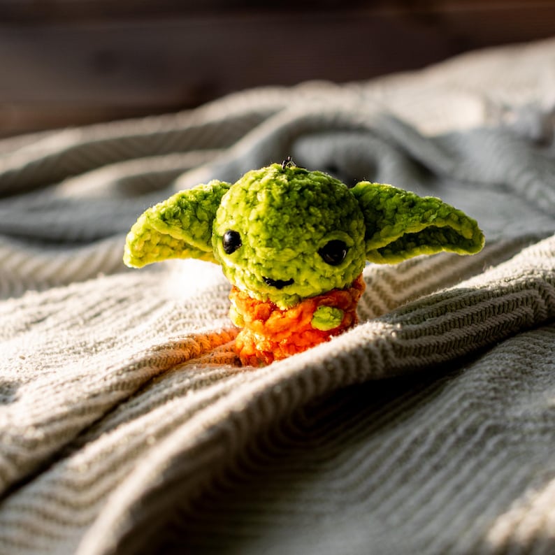 Puede incluir: Un peque&ntilde;o peluche de ganchillo hecho a mano con una cabeza redonda verde, ojos grandes, oscuros y brillantes, y orejas anchas y puntiagudas. El cuerpo est&aacute; texturizado con hilo naranja brillante, dando un aspecto suave y esponjoso a la figura.
