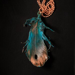 Puede incluir: Un collar de cadena de cobre con un colgante de pluma de color turquesa y cobre. La pluma tiene una textura detallada y un agujero en el centro.