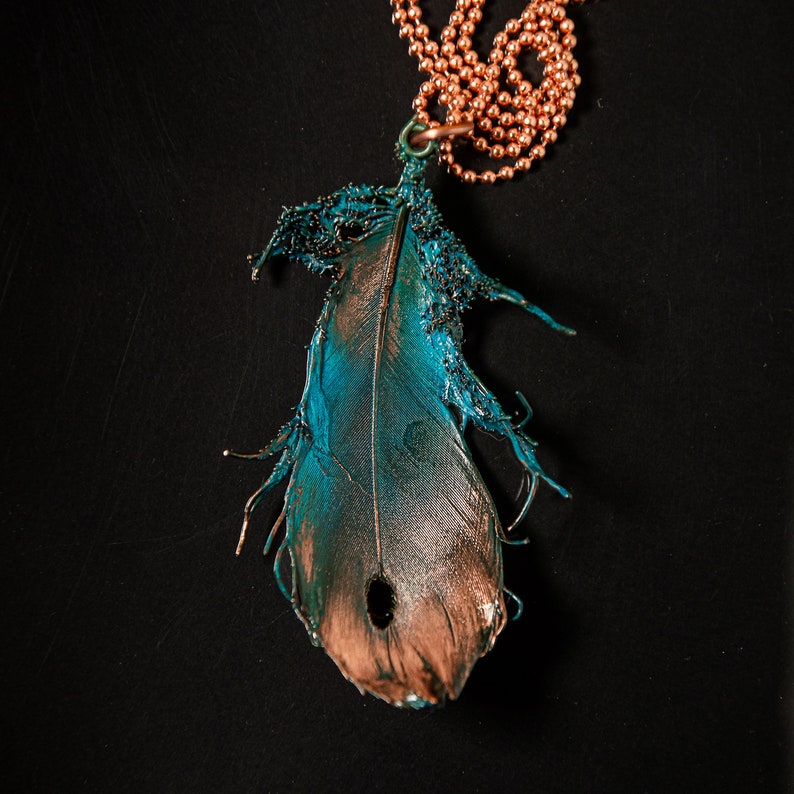 Puede incluir: Un collar de cadena de cobre con un colgante de pluma de color turquesa y cobre. La pluma tiene una textura detallada y un agujero en el centro.