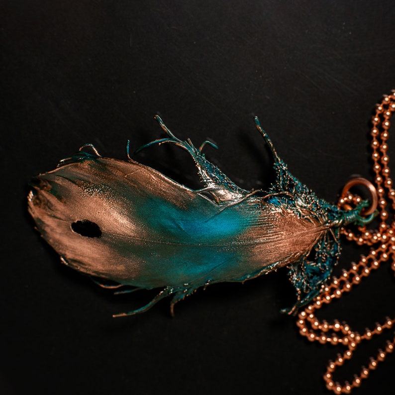 Puede incluir: Un collar con un colgante de pluma dorado y turquesa. La pluma est&aacute; detallada con una superficie texturizada y cuelga de una cadena dorada.