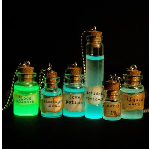 May include: Seven small glass bottles with cork stoppers and silver chains. The bottles are filled with glowing green and blue liquid. Each bottle has a label with a different inscription, including "blood unicorn", "love potion", "antidote covid-19", and "liquid luck".