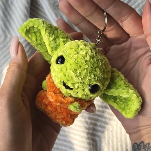 Puede incluir: Un peque&ntilde;o llavero de peluche tejido a mano que representa una criatura verde con orejas grandes y puntiagudas y ojos de pl&aacute;stico negro. La figura est&aacute; envuelta en una tela naranja texturizada y tiene una anilla de metal plateado.