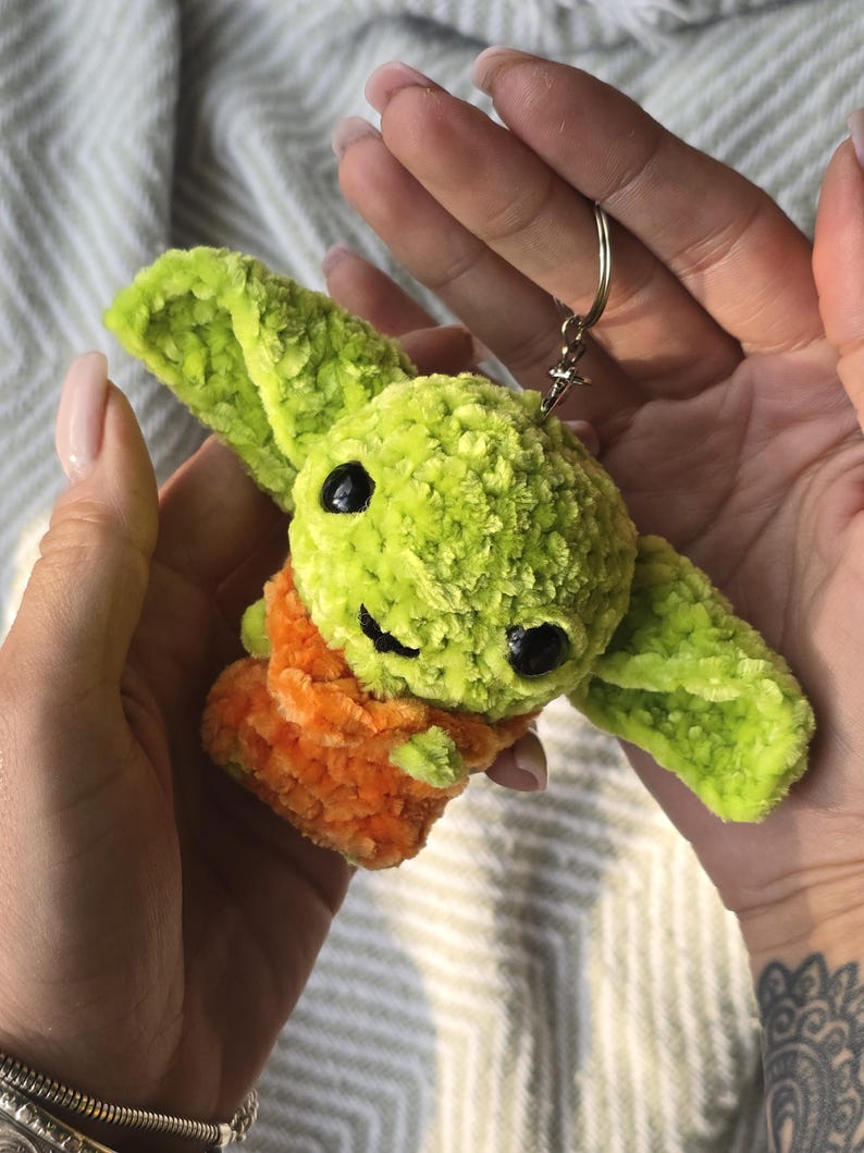 Puede incluir: Un peque&ntilde;o llavero de peluche tejido a mano que representa una criatura verde con orejas grandes y puntiagudas y ojos de pl&aacute;stico negro. La figura est&aacute; envuelta en una tela naranja texturizada y tiene una anilla de metal plateado.