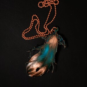 Puede incluir: Un collar de cadena de cobre con un colgante con forma de pluma. La pluma est&aacute; pintada en tonos de azul verdoso y dorado.