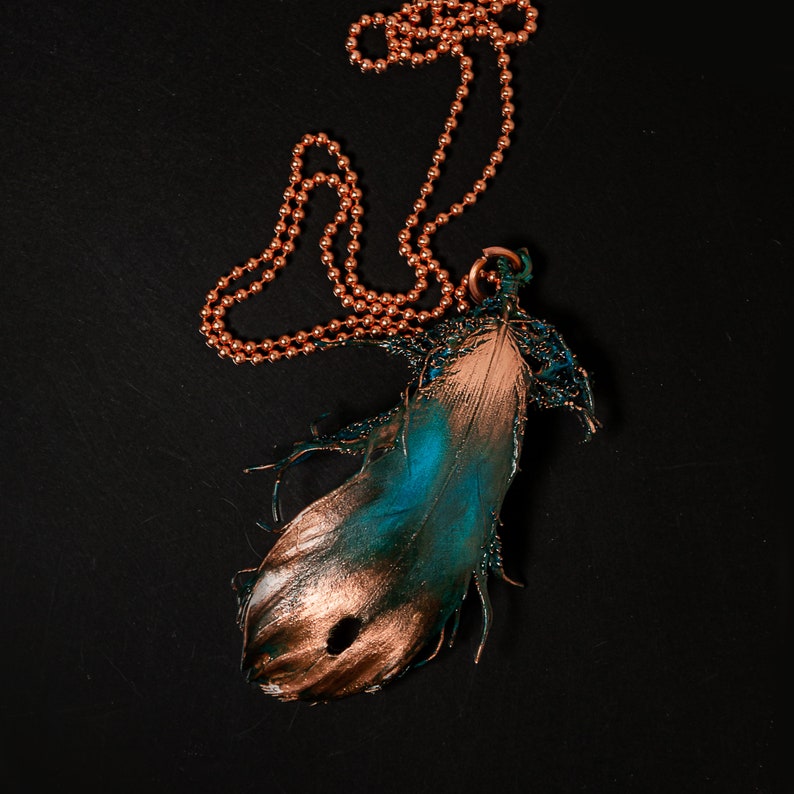 Puede incluir: Un collar de cadena de cobre con un colgante con forma de pluma. La pluma est&aacute; pintada en tonos de azul verdoso y dorado.