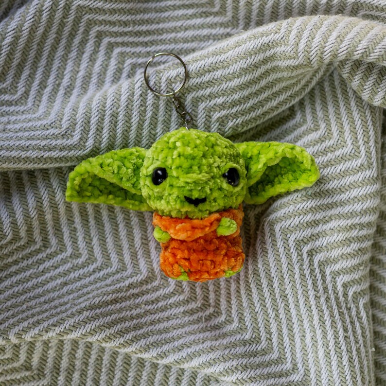 Puede incluir: Un llavero de ganchillo con forma de peque&ntilde;a criatura verde de orejas anchas, grandes ojos negros y una peque&ntilde;a sonrisa. La figura lleva una prenda naranja y est&aacute; sujeta a una anilla met&aacute;lica de color plateado en la parte superior.