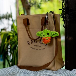 Puede incluir: Un bolso de lona color canela con asas marrones y el texto "BAG ART SINCE 1908 FASHION BAG FOR YOU" impreso en marr&oacute;n oscuro. Un peque&ntilde;o llavero de criatura de ganchillo verde y naranja con grandes ojos negros est&aacute; sujeto al herraje met&aacute;lico.