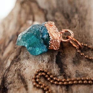 May include: A copper pendant necklace featuring a large, raw, teal blue crystal. The crystal is wrapped in copper and has a copper bail. The necklace is on a copper chain.