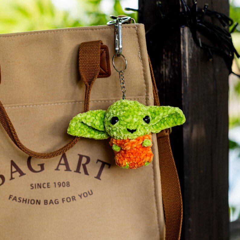 Puede incluir: Un bolso de lona color canela con correas marrones y el texto BAG ART SINCE 1908 FASHION BAG FOR YOU. Unido al bolso hay un llavero de peluche con forma de criatura verde de orejas grandes y cuerpo naranja, sujeto por una argolla de metal plateado.