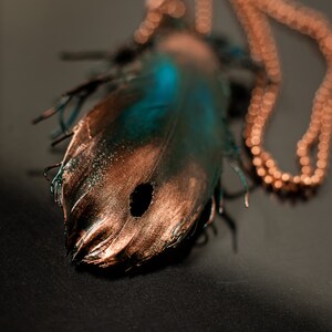 Puede incluir: Un colgante de pluma de cobre y turquesa en una cadena de cobre. La pluma tiene un agujero en el centro y est&aacute; detallada con un acabado texturizado.