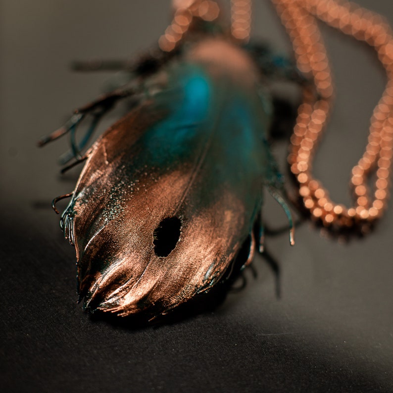 Puede incluir: Un colgante de pluma de cobre y turquesa en una cadena de cobre. La pluma tiene un agujero en el centro y est&aacute; detallada con un acabado texturizado.