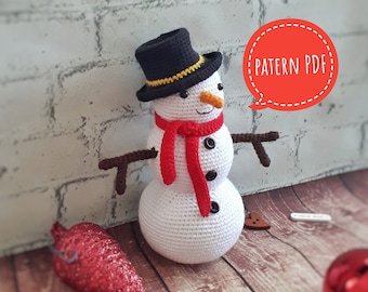 May include: A crocheted snowman wearing a black top hat, a red scarf, and twig arms. The snowman has a carrot nose and black button eyes. The image includes a red glittery pinecone and the text "PATTERN PDF".