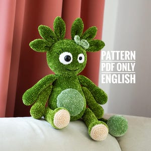 以下が含まれることがあります： 手作りの緑色の編みぐるみで、大きな白い目があり、明るい色の表面に座っています。 ぬいぐるみは丸いお腹、小さな腕、小さなリボンが付いています。 テキスト「PATTERN PDF ONLY ENGLISH」が表示されています。