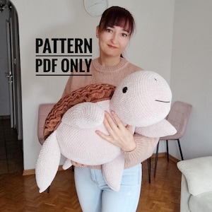 Puede incluir: Una gran tortuga de ganchillo rosa claro y suave con un caparazón marrón. La tortuga tiene una cara sonriente y está siendo sostenida. El texto "PATTERN PDF ONLY" se muestra en la imagen.