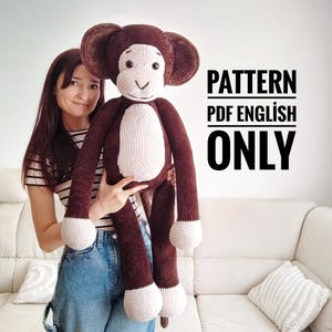 Könnte beinhalten: Ein großer, handgefertigter Häkelaffe in Braun- und Cremetönen. Der Affe hat große Ohren, einen weißen Bauch und cremefarbene Füße. Der Text "PATTERN PDF ENGLISH ONLY" ist in Schwarz dargestellt und weist darauf hin, dass eine Anleitung für den Affen verfügbar ist.