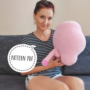 May include: A large, soft, pink crocheted pillow with a rounded body and a long, cylindrical appendage. The pillow is held in front of a person. The image includes a circular graphic with the text "PATTERN PDF".