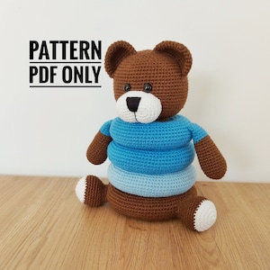Può includere: Un orsacchiotto all'uncinetto marrone fatto a mano con muso e zampe bianche, che indossa una camicia a righe blu. L'orso è seduto su una superficie di legno. Il testo "PATTERN PDF ONLY" è nell'immagine.