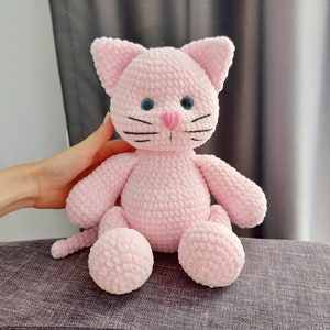May include: A pink crochet cat toy with blue eyes, a pink nose, and black whiskers. The cat has a soft, textured appearance and is sitting upright. The plush toy is a cute and cuddly item.