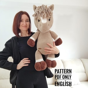 Op de afbeelding: Een groot, handgemaakt gehaakt paard in tinten beige, bruin en wit. Het paard heeft een pluizige manen en staart, met bruine hoeven en een witte bles op het gezicht. De tekst "PATTERN PDF ONLY ENGLISH" is zichtbaar.