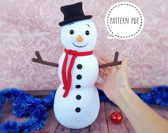 May include: A handmade white crochet snowman with a black top hat, a red scarf, and brown twig arms. The snowman has black button eyes and a carrot nose. The text "PATTERN PDF" is in a speech bubble.