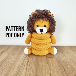 Peut inclure: Un jouet lion en crochet avec une crinière marron et un corps jaune. Le lion a un museau et des pattes blancs. Le texte "PATTERN PDF ONLY" est affiché en noir. Le jouet est posé sur une surface en bois.