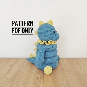 May include: A handmade blue crochet dinosaur toy with yellow detailing. The dinosaur features a ruffled collar and a stackable body. The text "PATTERN PDF ONLY" is displayed in black and white.
