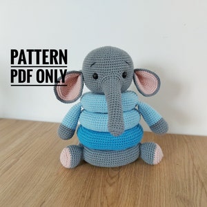 May include: A crocheted elephant toy with a stack of blue rings. The elephant is grey with pink inner ears and feet. The text "PATTERN PDF ONLY" is displayed in black.