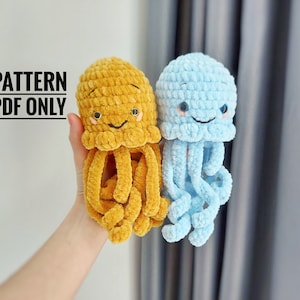 May include: Two crocheted jellyfish toys, one mustard yellow and one light blue. They have smiling faces with embroidered eyes and tentacles. The text "PATTERN PDF ONLY" is above the toys.