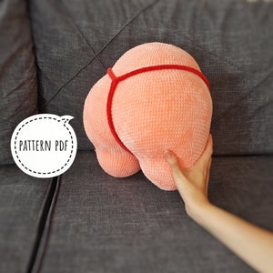 May include: A peach-coloured, plush, crocheted cushion shaped like a bottom, with a red strap detail. The cushion is held by a hand. The text "PATTERN PDF" is in a white circle.