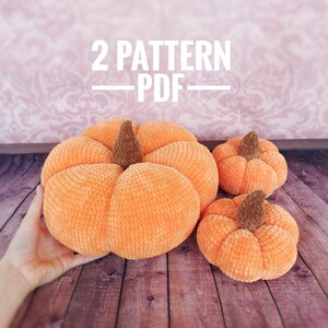 May include: Three plush orange pumpkins with brown stems are displayed on a wooden surface. The largest pumpkin is being held by a hand. The text "2 PATTERN PDF" is displayed in white across the top of the image.