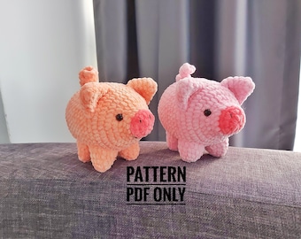 May include: Two plush, crocheted pig figurines. One is peach-colored, the other is pink. Both have pink snouts, small black eyes, and small ears. The text "PATTERN PDF ONLY" is printed below the pigs.
